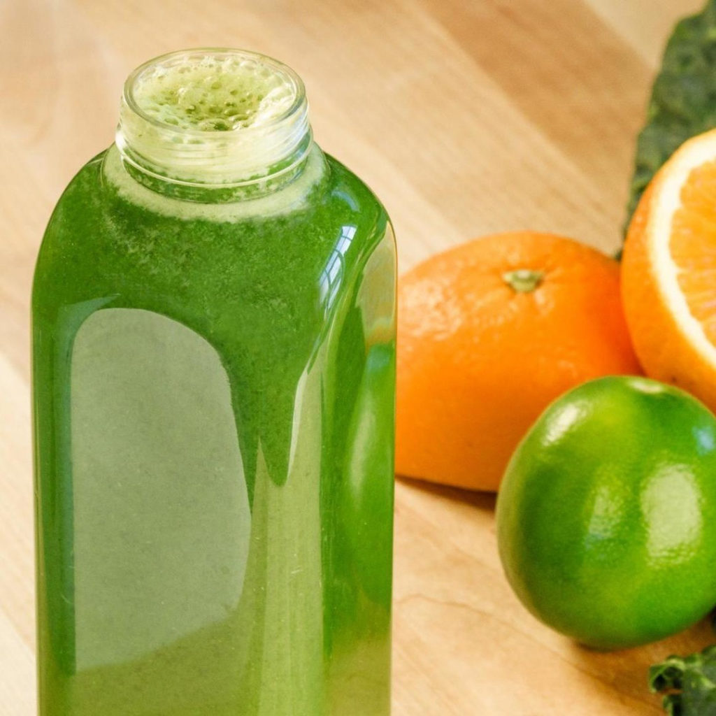 Photo of a bottle of green juice and lime and oranges in the background.