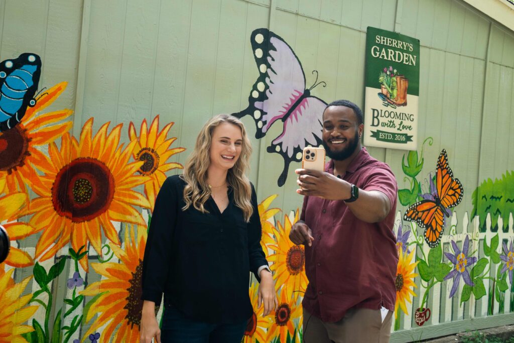 Photo of two people taking a selfie infant of a mural with butterflies and sunflowers. 