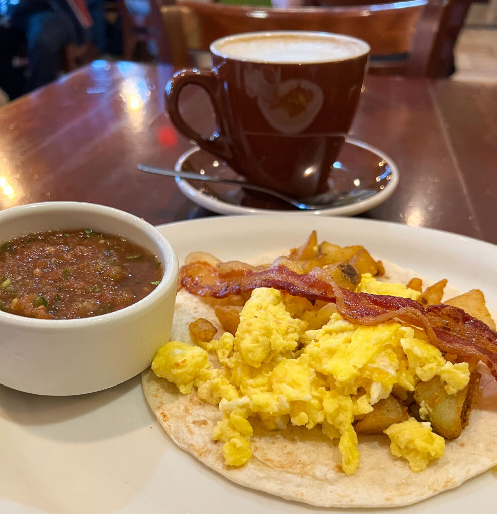 Photo of Breakfast taco with bacon, egg, potato and salsa and a coffee cup in the background. 